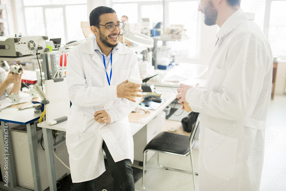 Happy excited young Arabian lab technician with badge on neck laughing at colleague story while they enjoying coffee break in manometer plant