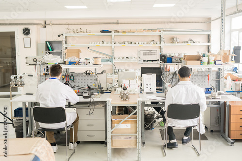 Rear view of busy engineers in lab coats sitting on chairs at table and assembling or repairing measuring device in manometer workshop