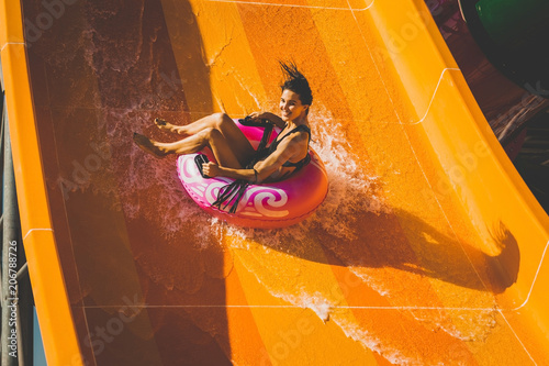 pretty brunette slim woman on the rubber ring having fun going down on the orange water slide in the aqua park. Summer Vacation. Weekend on resort