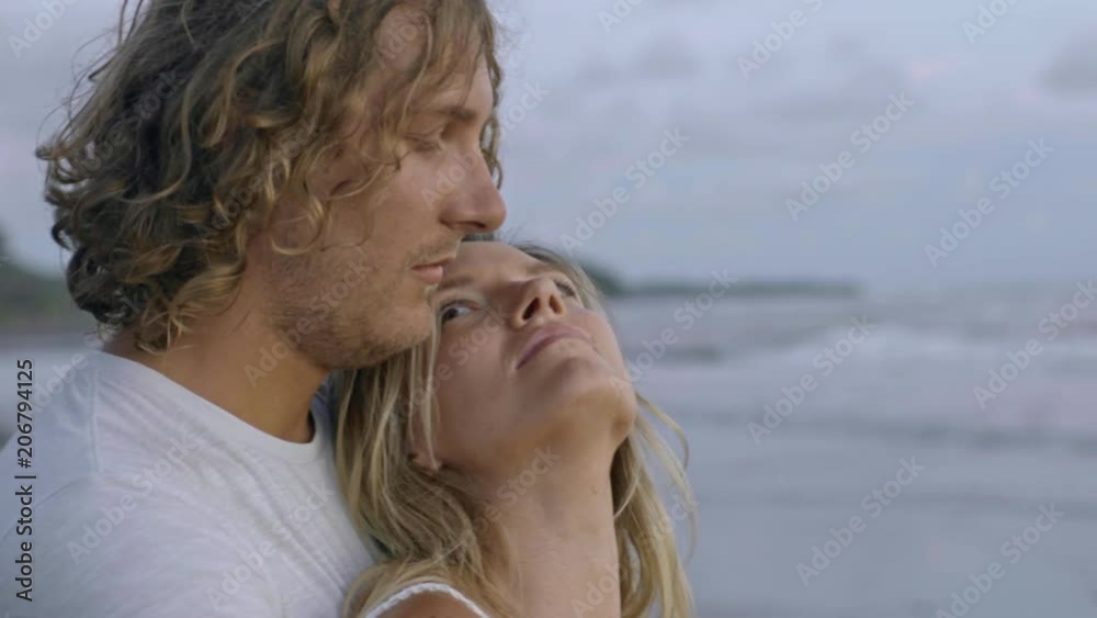 Handheld PAN of young man with curly hair hugging and kissing his girlfriend while standing on beach and looking at ocean Handheld PAN of young man with curly hair hugging and kissing his girlfriend while standing on beach and looking at ocean