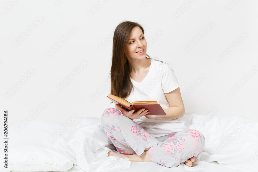 Young happy woman sitting in bed with white sheet, pillow, blanket on white background. Beauty female spending time in room, reading book, enjoying of rest. Relax, good mood concept. Place for text.