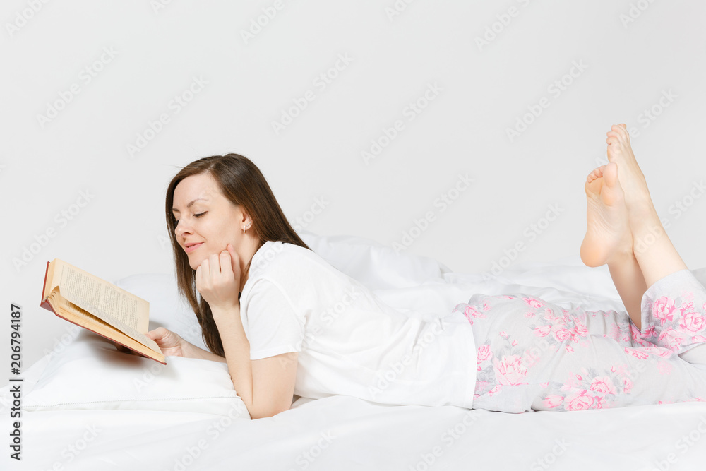 Young happy woman lying in bed with white sheet, pillow, blanket on white background. Beauty female spending time in room, reading book, enjoying of rest. Relax, good mood concept. Place for text.