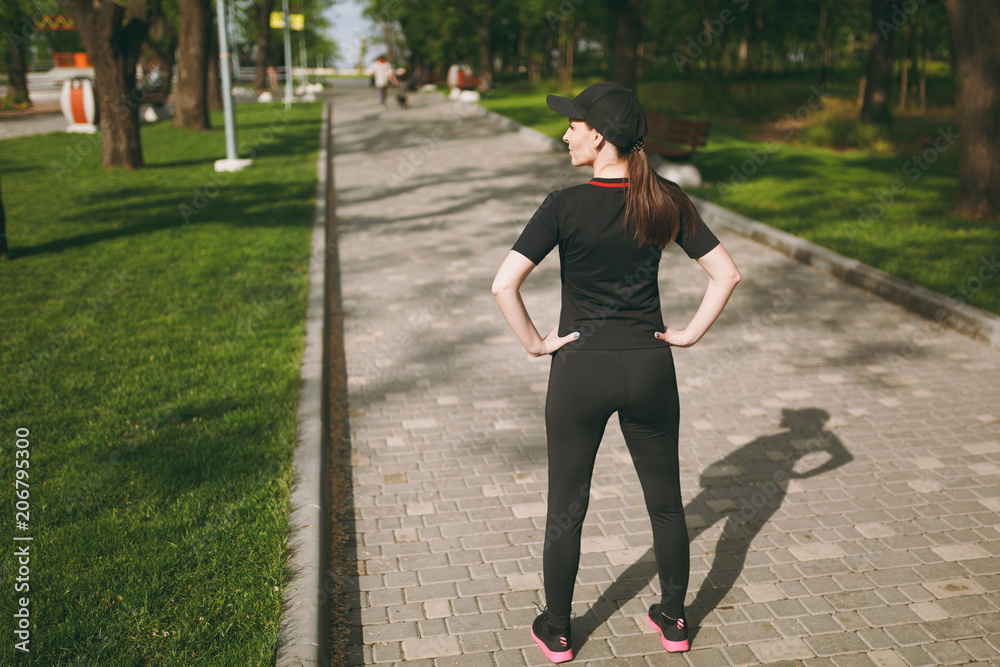 Back view Young athletic beautiful brunette girl in black uniform, cap standing, resting and looking aside before or after running, training in city park outdoors. Fitness, healthy lifestyle concept. Back view Young athletic beautiful brunette girl in black uniform, cap standing, resting and looking aside before or after running, training in city park outdoors. Fitness, healthy lifestyle concept.