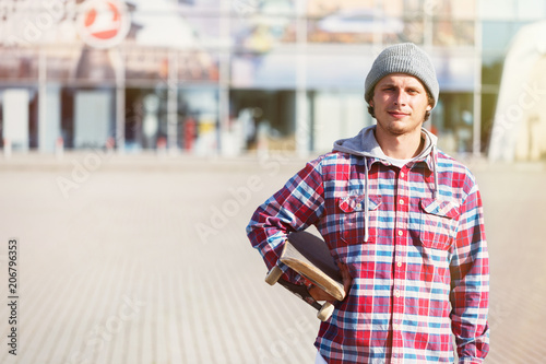 Portrait of hipster man dressed in cheked shirt and cap holding skateboard before modern building photo