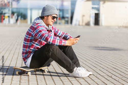Portrait of hipster man with sunglasses holding smartphone sitting on the skateboard