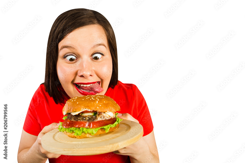 Young fat woman holds hamburger, smiles with his tongue hanging out puts out his tongue isolated on light pink background.