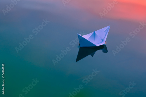 Paper origami boat floats on the surface of the water at sunset or dawn