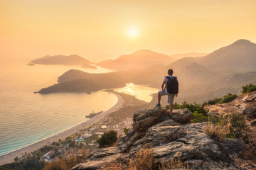 Young sporty man with backpack standing on the top of rock and looking at the seashore and mountains at sunset in summer. Scene with man, sea, mountain ridges and orange sky with sun. Oludeniz, Turkey