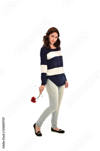 full length portrait of girl wearing striped blue and white jumper and jeans, holding a red flower. standing pose on white studio background