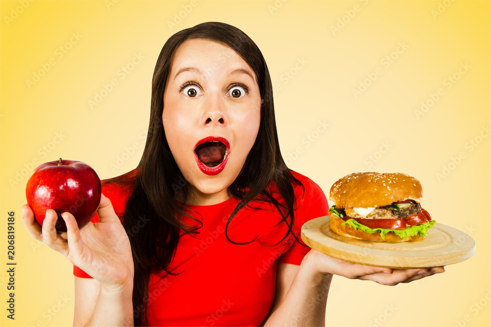 Portrait of a beautiful fat girl in surprise and holding a red apple and a hamburger, isolated on yellow background . Portrait of a beautiful fat girl in surprise and holding a red apple and a hamburger, isolated on yellow background .