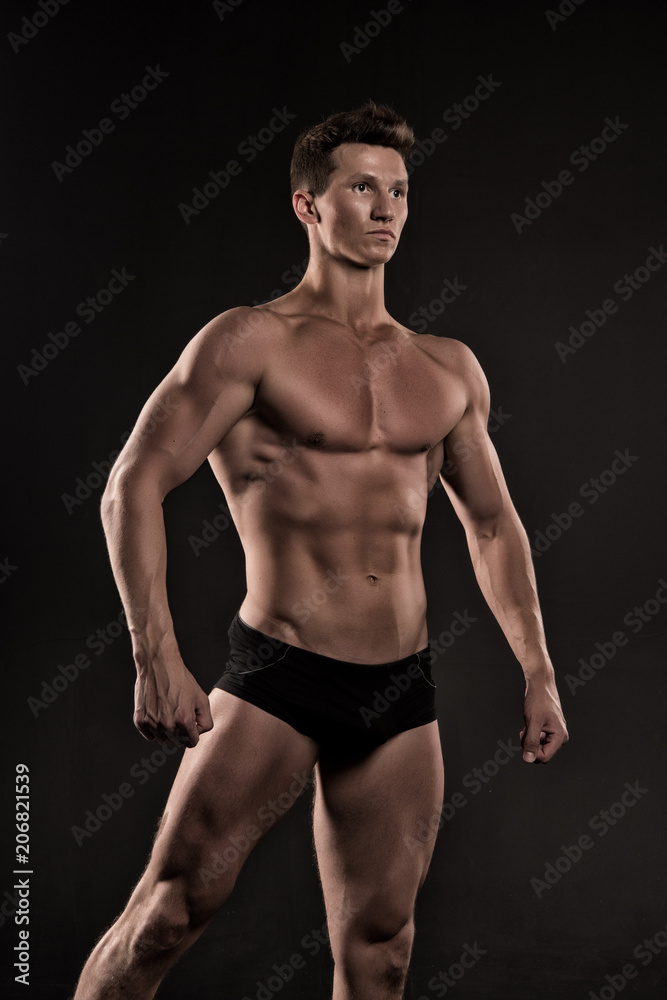 Macho with muscular body posing as bodybuilder, black background. Bodybuilder in black underpants shows his muscles. Athlete on confident face demonstrates muscular body. Bodybuilding concept