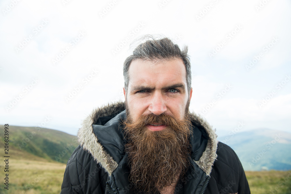 Man with long beard and mustache wears jacket. Hipster on strict face with beard looks brutally while hiking. Hermit concept. Man with brutal bearded appearance, brutal unshaven man looks untidy