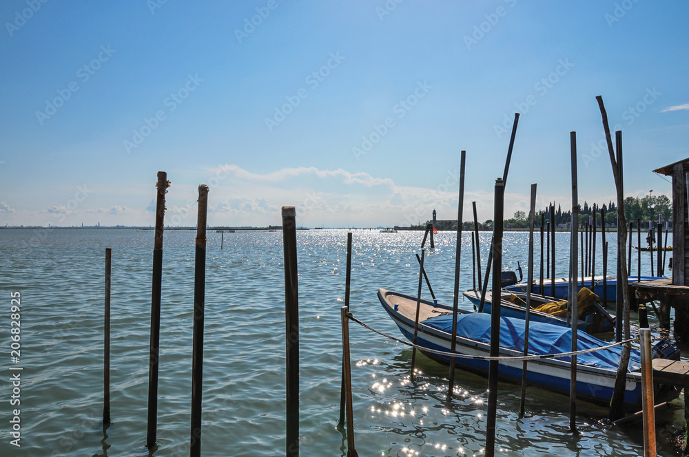 Panoramic view of Venice lagoon with pier and boats with blue and sunny sky in Burano, a gracious little town full of canals, near Venice. Located in the Veneto region, northern Italy