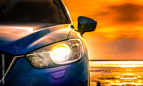 Blue compact SUV car with sport and modern design parked on concrete road by the sea at sunset. Environmentally friendly technology. Business success concept. Car with open headlamp light. © Artinun