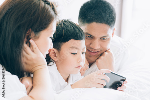 Asian boy playing smartphone with young parents lying in bed.Happy famility