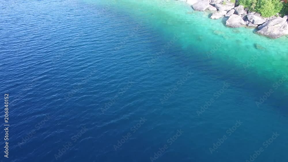 Drone flying above bright blue water slowly reveals and flies over cliffs Drone flying above bright blue water slowly reveals and flies over cliffs