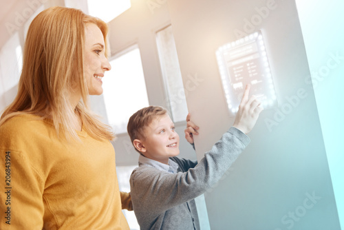 Happy relatives. Positive curious boy standing next to his kind smiling mother and carefully touching a transparent security system on the wall