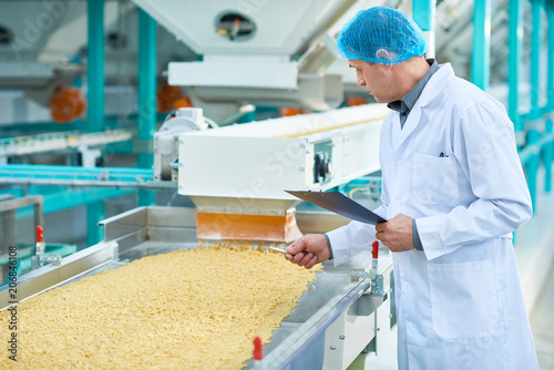 Side view portrait of senior factory worker doing  production quality inspection in food industry holding clipboard standing by conveyor belt, copy space