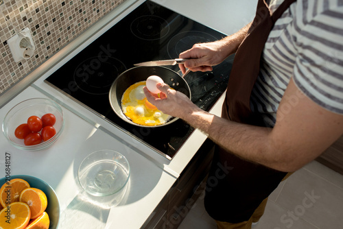 Close-up of unrecognizable man in apron cracking egg with knife while cooking fried eggs in cooking pan in domestic kitchen