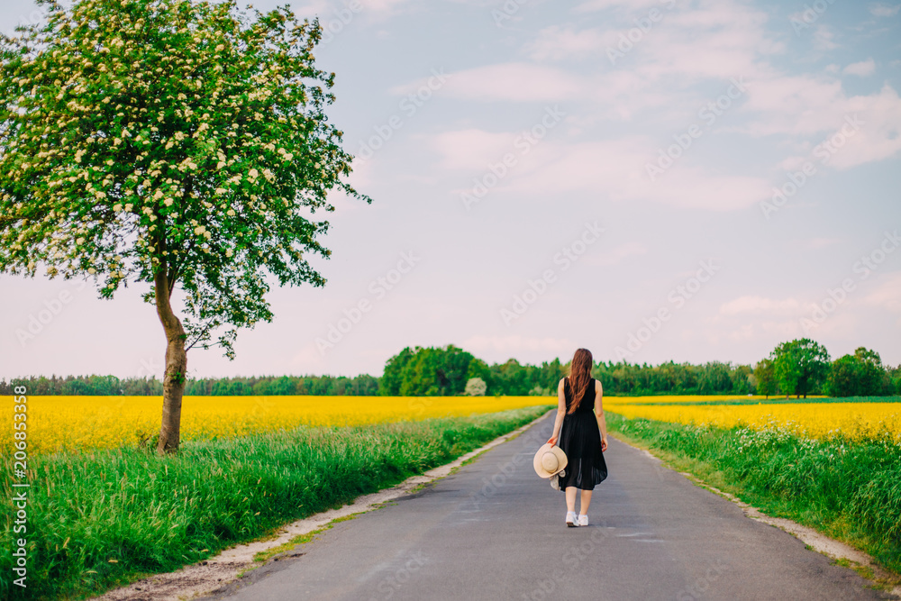 Fototapeta premium A girl walking the road near rapeseed field