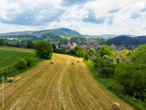 Straw bales on the field near Saschiz fortified church in Saschiz villages, Sibiu, Transylvania, Romania. Agriculture landscape, green field and clouds on blue sky. A green meadow with rolled up wheat photo