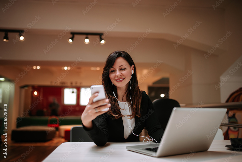 Image of smiling woman with earphones texting on smartphone while sitting in front of laptop Image of smiling woman with earphones texting on smartphone while sitting in front of laptop