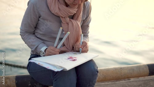 a young woman holds colored pens in her hands and draws flowers, while waiting for her friends, she is near the water in the cool autumn season photo