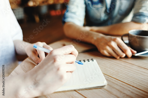 Hands of woman making notes in her notepad while discussing business ideas with her male partner over coffee in cafe