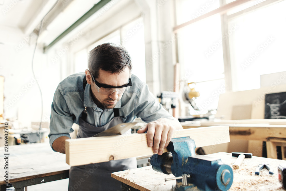 Portrait of carpenter in protective eyewear sawing carefully wood plank with hand saw in his workshop Portrait of carpenter in protective eyewear sawing carefully wood plank with hand saw in his workshop