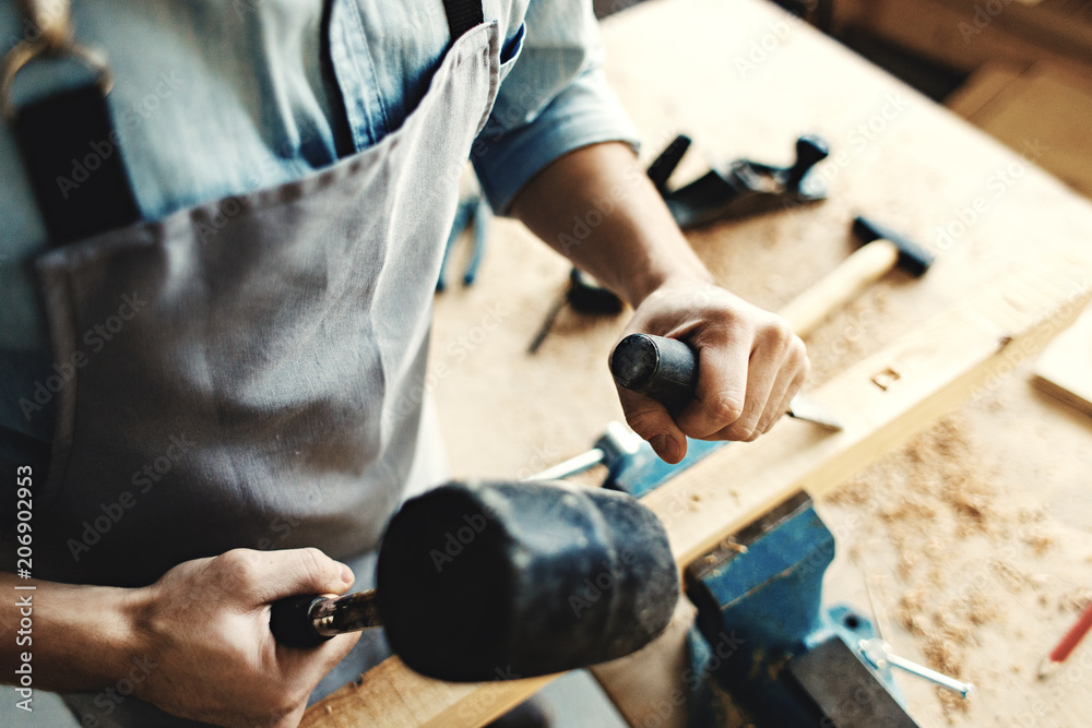 Hands of skillful woodworker using hammer and chisel to make mortise in wood plank, high angle view.