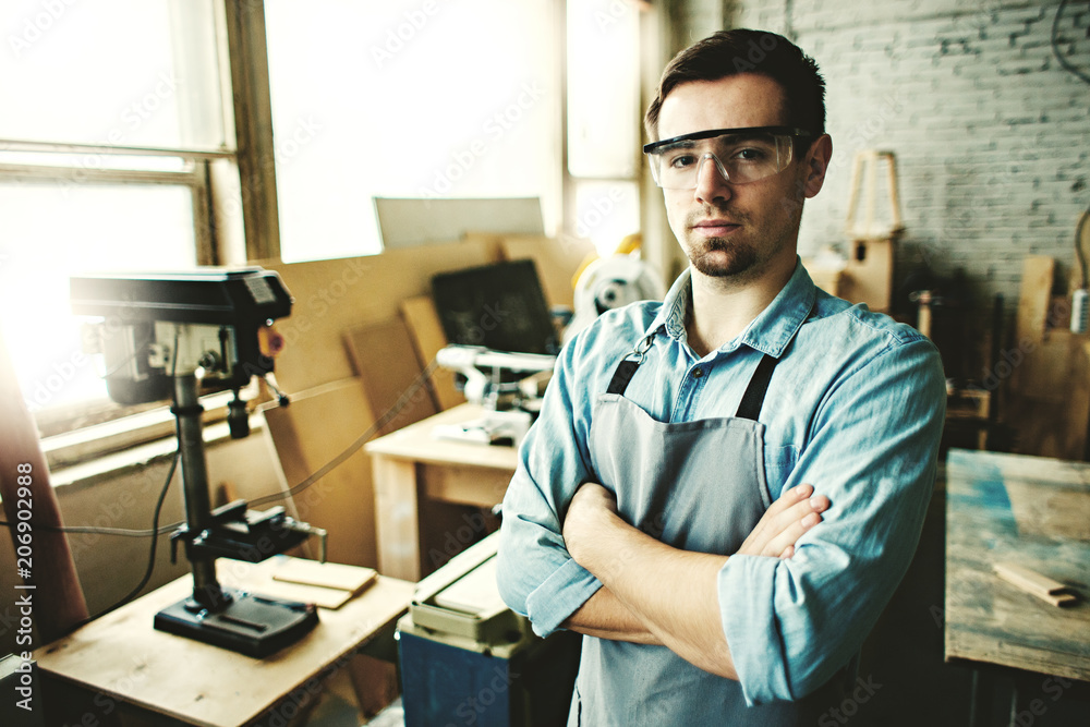 Portrait of confident young carpenter wearing apron and protective eyeglasses posing cross-armed in his woodworking shop. Portrait of confident young carpenter wearing apron and protective eyeglasses posing cross-armed in his woodworking shop.