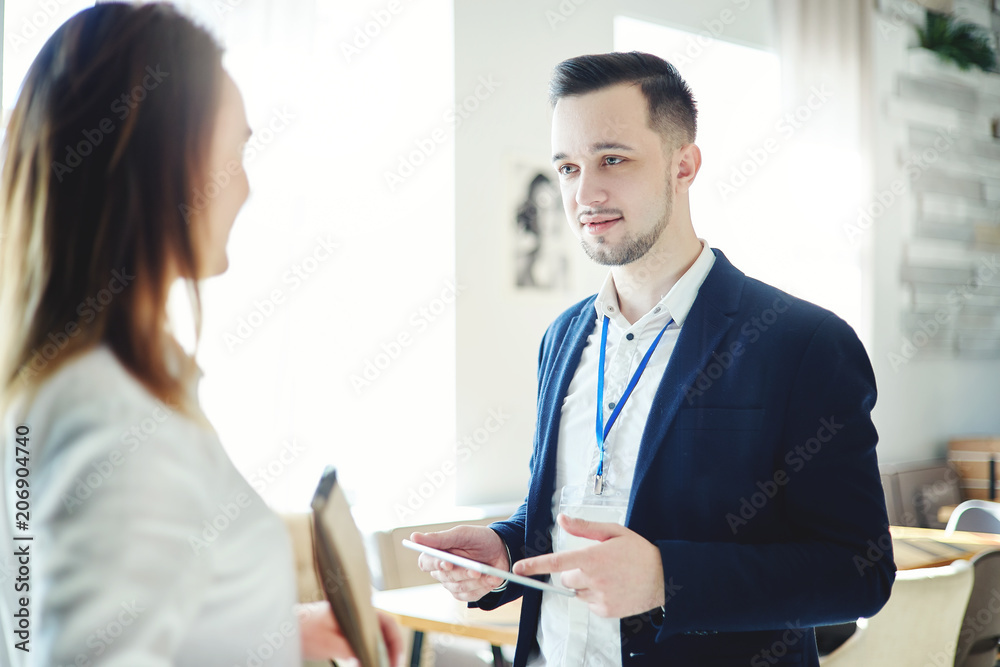 Confident young businessman with tablet computer in his hands and accreditation badge around his neck talking with unrecognizable female colleague at business event Confident young businessman with tablet computer in his hands and accreditation badge around his neck talking with unrecognizable female colleague at business event