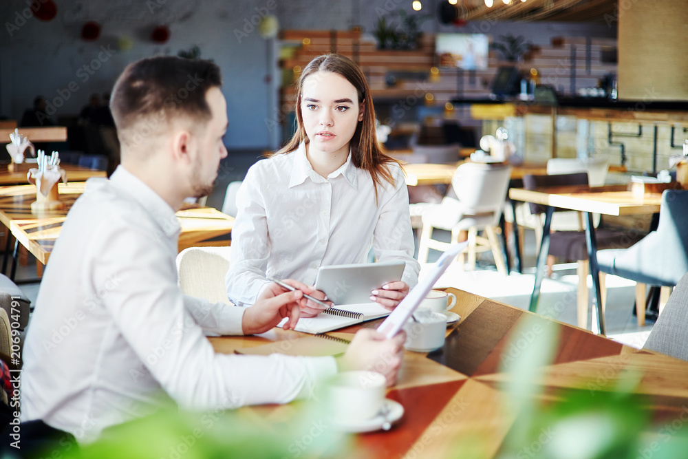 Young business people discussing project at cafe. Businessman with documents in his hands and businesswoman with tablet computer sitting at table and talking Young business people discussing project at cafe. Businessman with documents in his hands and businesswoman with tablet computer sitting at table and talking