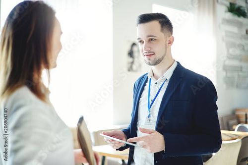 Confident young businessman with tablet computer in his hands and accreditation badge around his neck talking with unrecognizable female colleague at business event