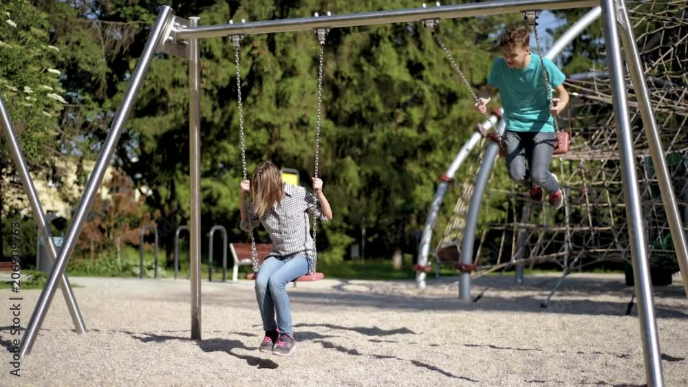 Happy two children ride on a swing at summer park. Cute teen boy and girl swings at playground outdoors. Happy two children ride on a swing at summer park. Cute teen boy and girl swings at playground outdoors.