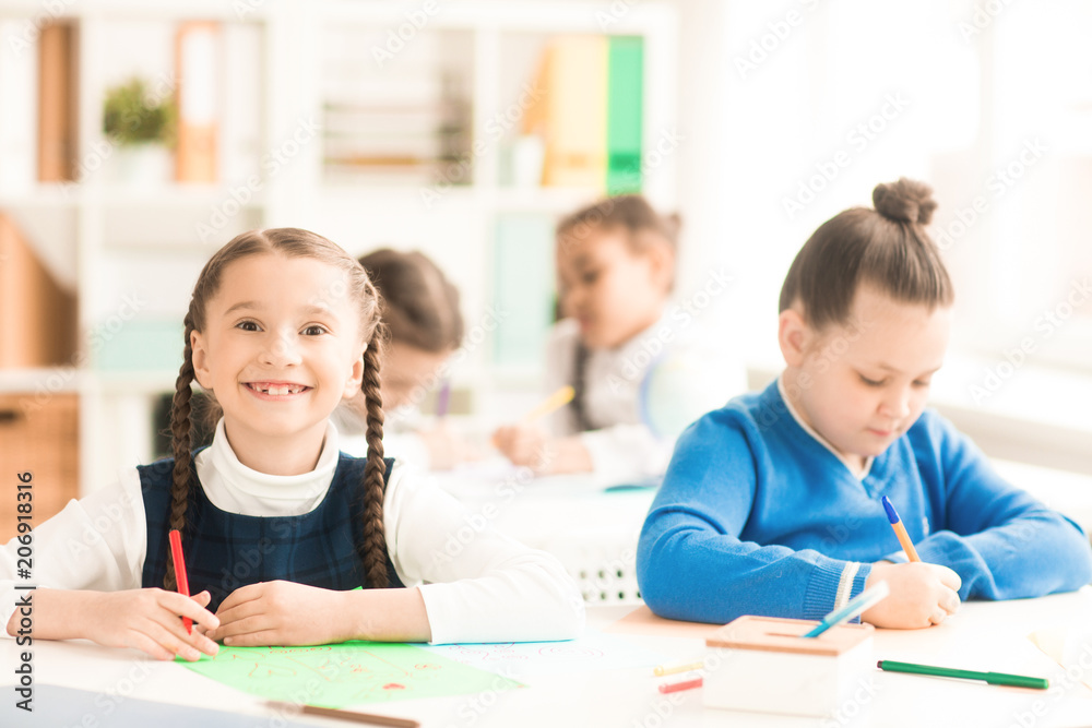 Portrait of smiling schoolgirl sitting at the desk with her classmte and drawing Portrait of smiling schoolgirl sitting at the desk with her classmte and drawing