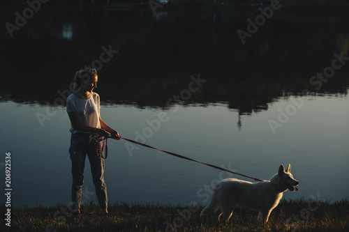 Side view of a long haired caucasian girl walking in the park around the lake with her white siberian husky at the sunset.