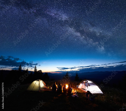 Family tourists mother, father, two guys resting at night camping in mountains, sitting on log beside campfire and two illuminated tents, enjoying amazing view of evening sky full of stars, Milky way
