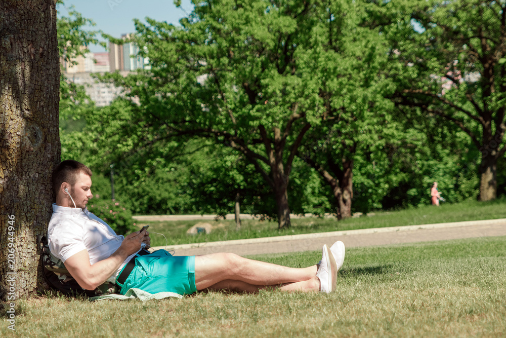 Young successful businessman in a white shirt. A man is sitting on the grass, working on a smartphone in a city park on a green lawn outdoors in nature. Mobile office, business concept