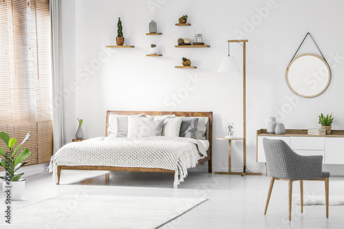 Front view of a bright natural bedroom interior with wooden bed with cover and pillows, nightstand and lamp, gray armchair and round mirror on white wall. Real photo.