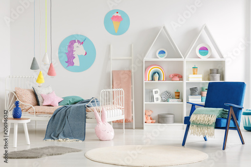 Colorful kid's bedroom interior with a unicorn and ice cream poster, bed with sheets, rabbit pillow, shelves and blue armchair with a blanket