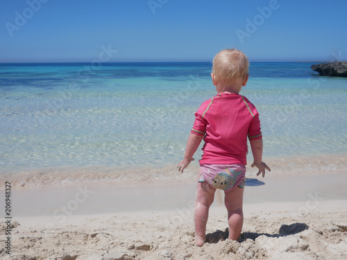 Little baby in pink swimming suit looking at the aquamarine blue see from a sand beach of Favignana island (Sicily, Italy).