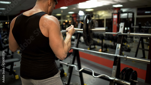 Sporty guy doing lift-ups with curl bar near stand in gym, pumping arm muscles