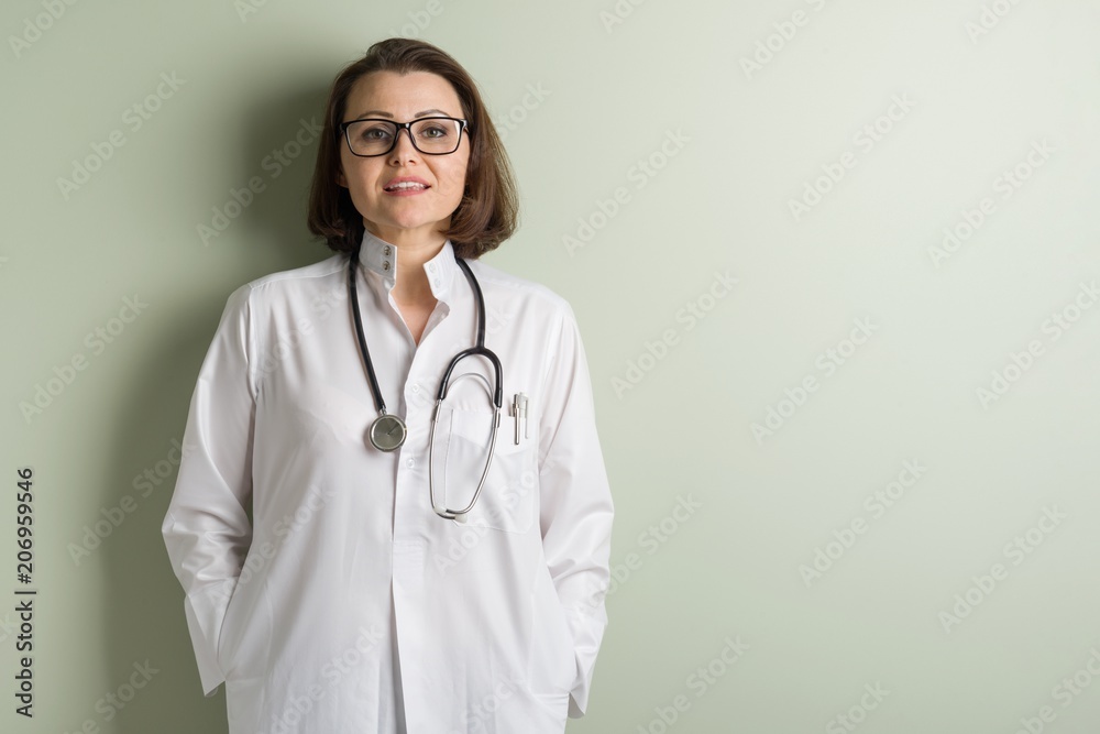 Positive middle-aged woman doctor, wearing glasses. Background green wall in the clinic. Positive middle-aged woman doctor, wearing glasses. Background green wall in the clinic.