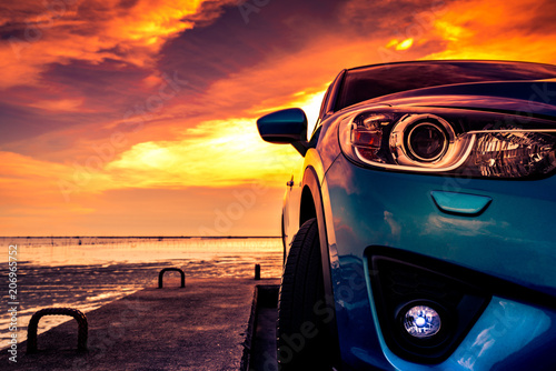 Blue compact SUV car with sport and modern design parked on concrete road by the sea at sunset. Environmentally friendly technology. Business success concept. Front view of car with open fog lamp © Artinun