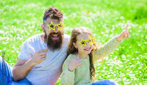 Rock star concept. Dad and daughter sits on grass at grassplot, green background. Family spend leisure outdoors. Child and father posing with star shaped eyeglases photo booth attribute at meadow. photo