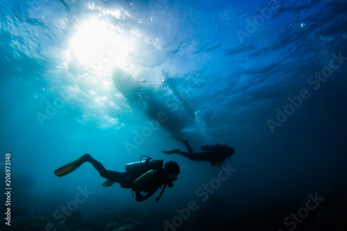 Two divers swim underwater in a tropical sea with a silhouette of a boat over them. Philippines