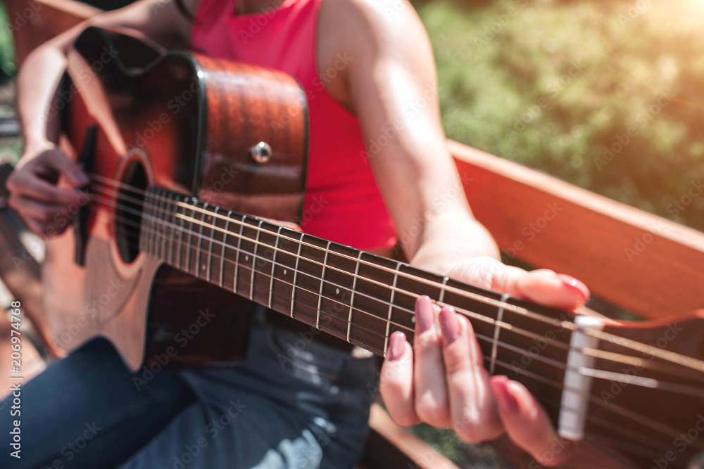 Close up of musician's hands playing the guitar. Girl is holding her fingers on guitar's strings. Girl is sitting on bench outside in this beautiful day.