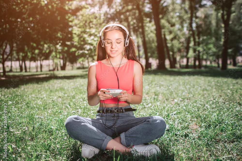 Peaceful and relaxed girl is sitting on grass with her legs crossed and listening to music. She has headphones on her ears. Girl is looking to music player and smiling. Peaceful and relaxed girl is sitting on grass with her legs crossed and listening to music. She has headphones on her ears. Girl is looking to music player and smiling.