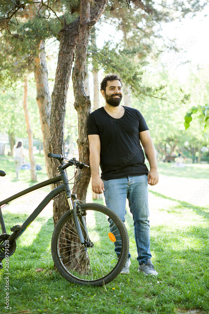 Smiling bearded man leaning on a tree in the park next to his bicycle